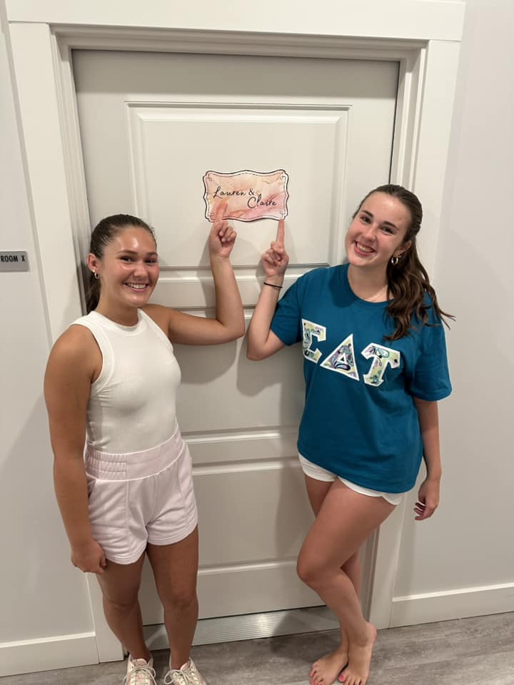 Two women smiling, pointing to a sign on a door bearing their names.